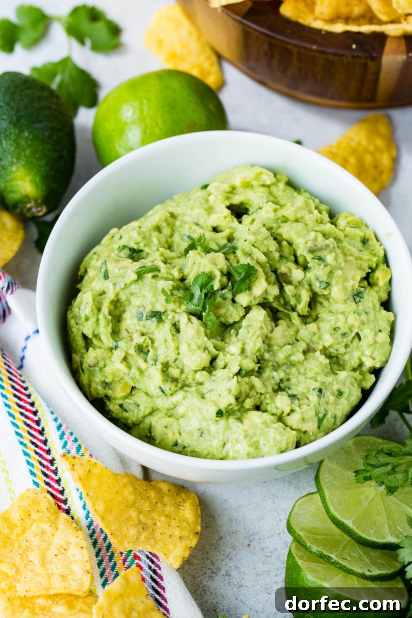 A vibrant bowl of homemade authentic guacamole, topped with fresh cilantro leaves. Two whole limes and a wooden bowl of golden tortilla chips are artfully arranged in the background, while lime slices and more chips are in front, creating an inviting tableau.