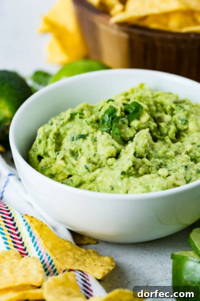 A pristine white bowl of fresh guacamole, generously topped with cilantro leaves. In the background, two whole limes and a rustic wooden bowl brimming with tortilla chips set a festive scene. More chips and bright lime slices are arranged in the foreground, highlighting the dip's vibrant appeal.