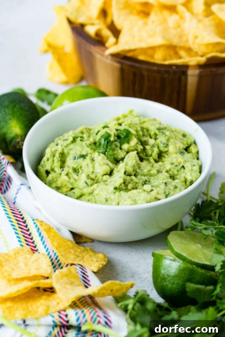 A captivating overhead view of a bowl of fresh guacamole, elegantly garnished with a few cilantro leaves. Scattered tortilla chips surround the bowl, alongside a partially sliced lime, hinting at the dip's zesty flavor. In the background, two whole limes and a wooden bowl of more tortilla chips complete the inviting scene.
