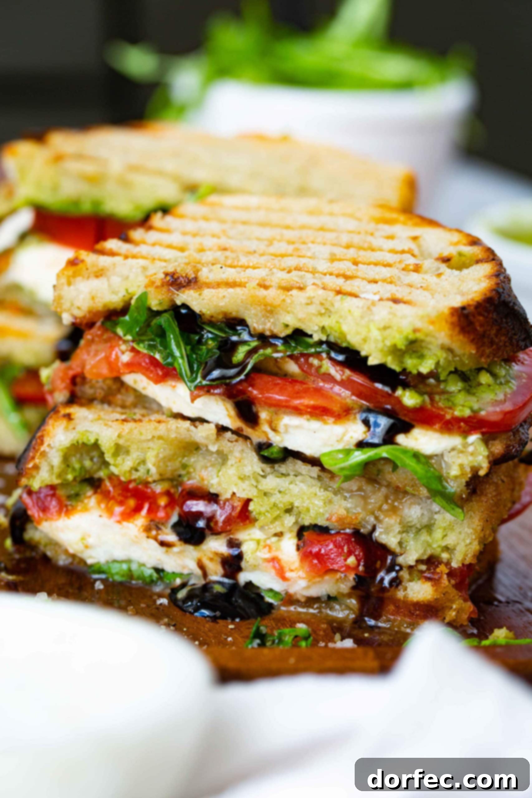 A balsamic glazed caprese sandwich that has been cut in half and stacked on top of each other. The sandwich has grill marks on top and has arugula, fresh mozzarella cheese slices and tomato slices inside. You can see the balsamic glaze dribbling out of the sandwich and another sandwich in the background.