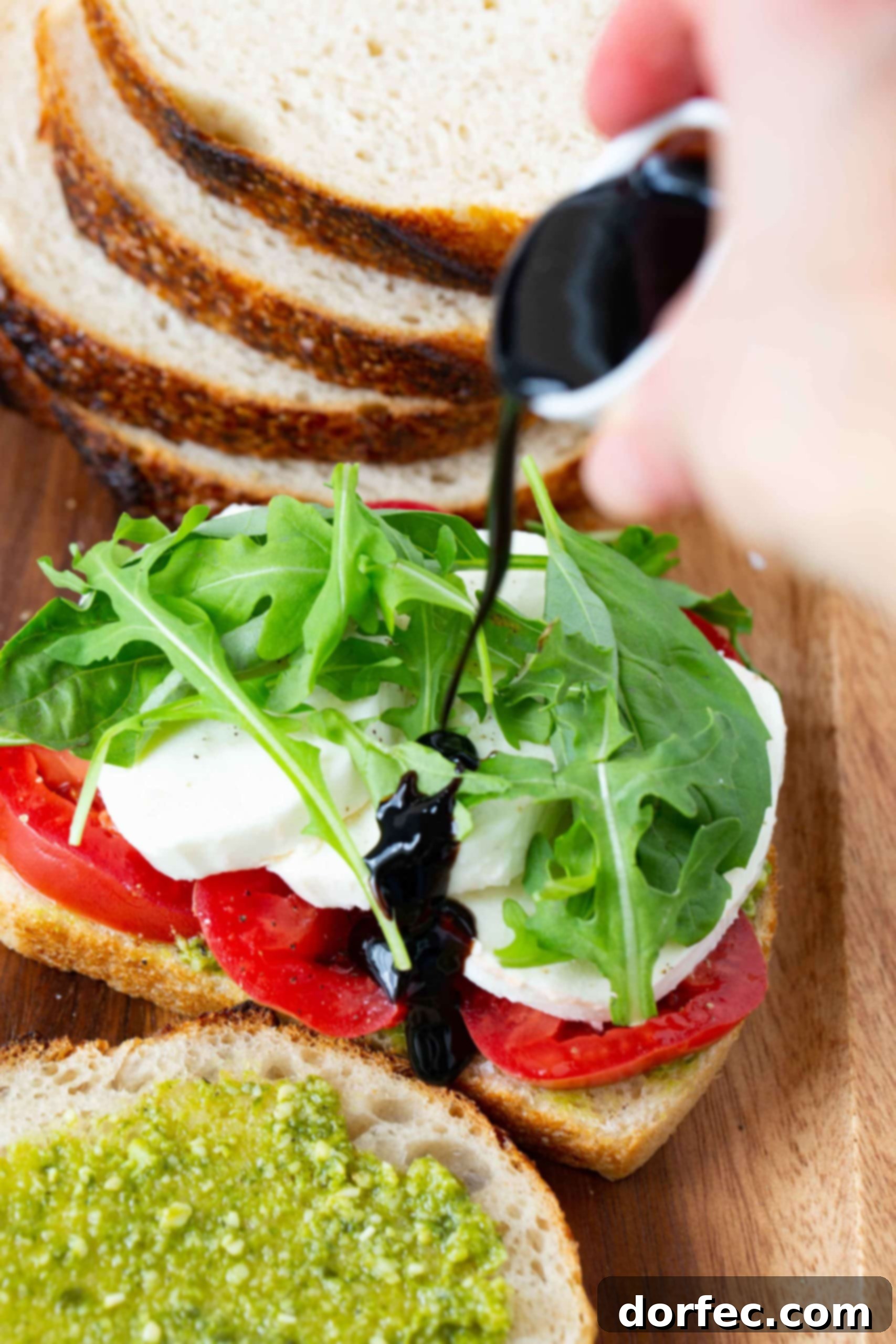 An opened sandwich with sliced tomatoes, sliced fresh mozzarella cheese, and arugula. A balsamic glaze is being drizzled over the top. There is another slice of bread that has been spread with pesto in front of it. there are four slices of bread stacked in the background.
