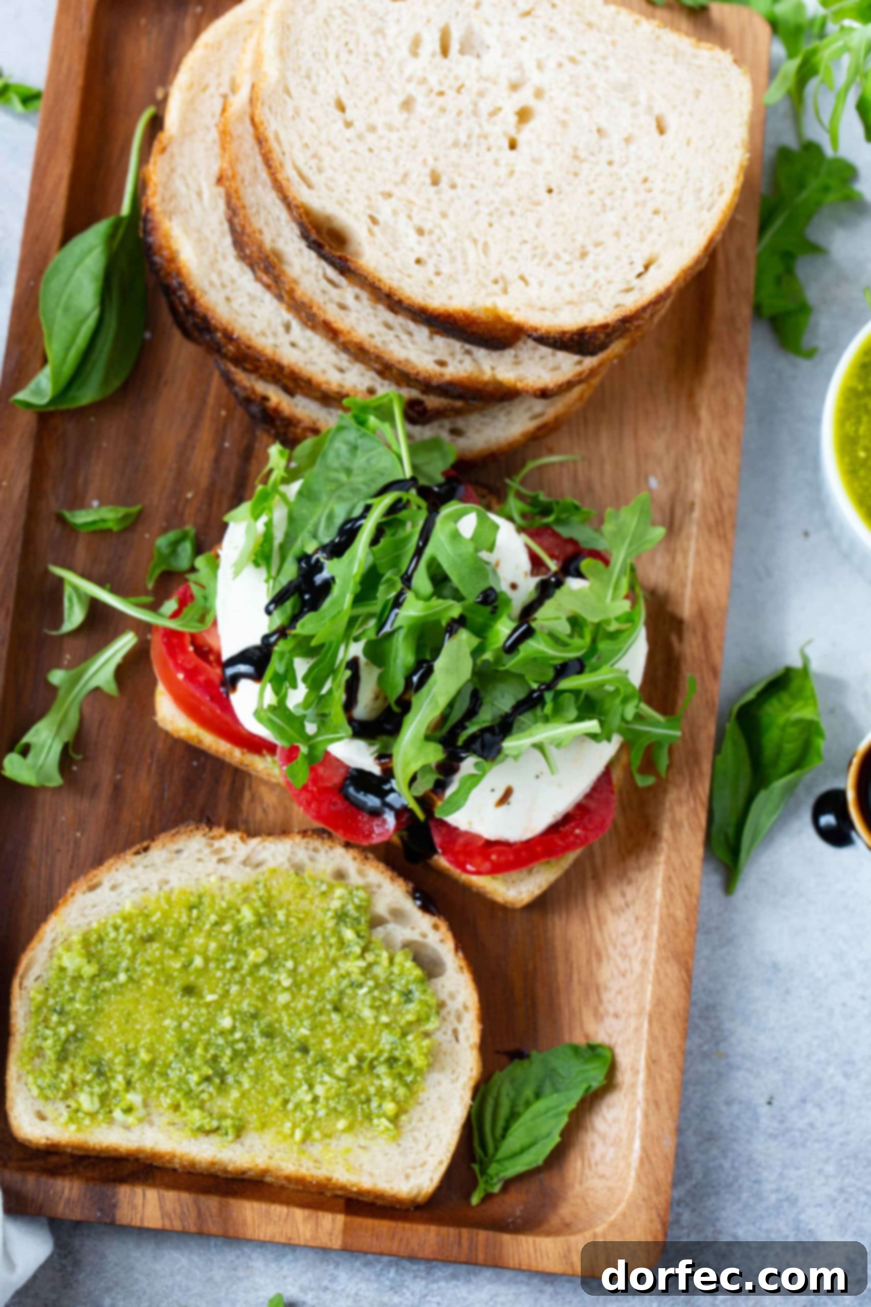 A wooden tray with sliced bread, a slice of bread with tomatoes, mozzarella cheese, and arugula on top. There is another slice of bread laying next to it that has been spread with pesto in front of it. Arugula and basil leaves lay on the tray and table next to the tray.