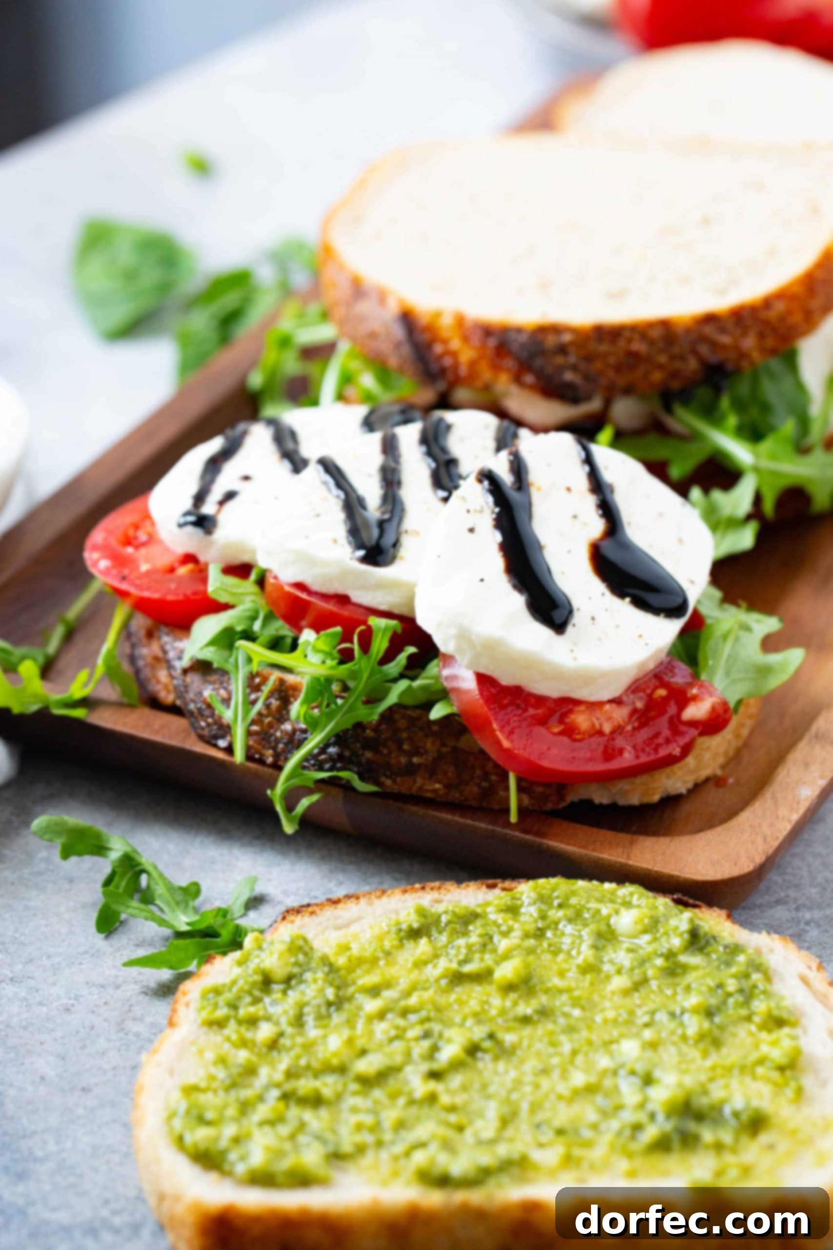 A slice of bread on a wooden tray. There are sliced tomatoes and fresh sliced mozzarella cheese and arugula on the slice of bread. A balsamic glaze is drizzled over the cheese. There are other slices of bread stacked on the tray next to the sandwich and arugula leaves scattered around. Another slice of bread is on the table in front of the tray and it has been spread with pesto.