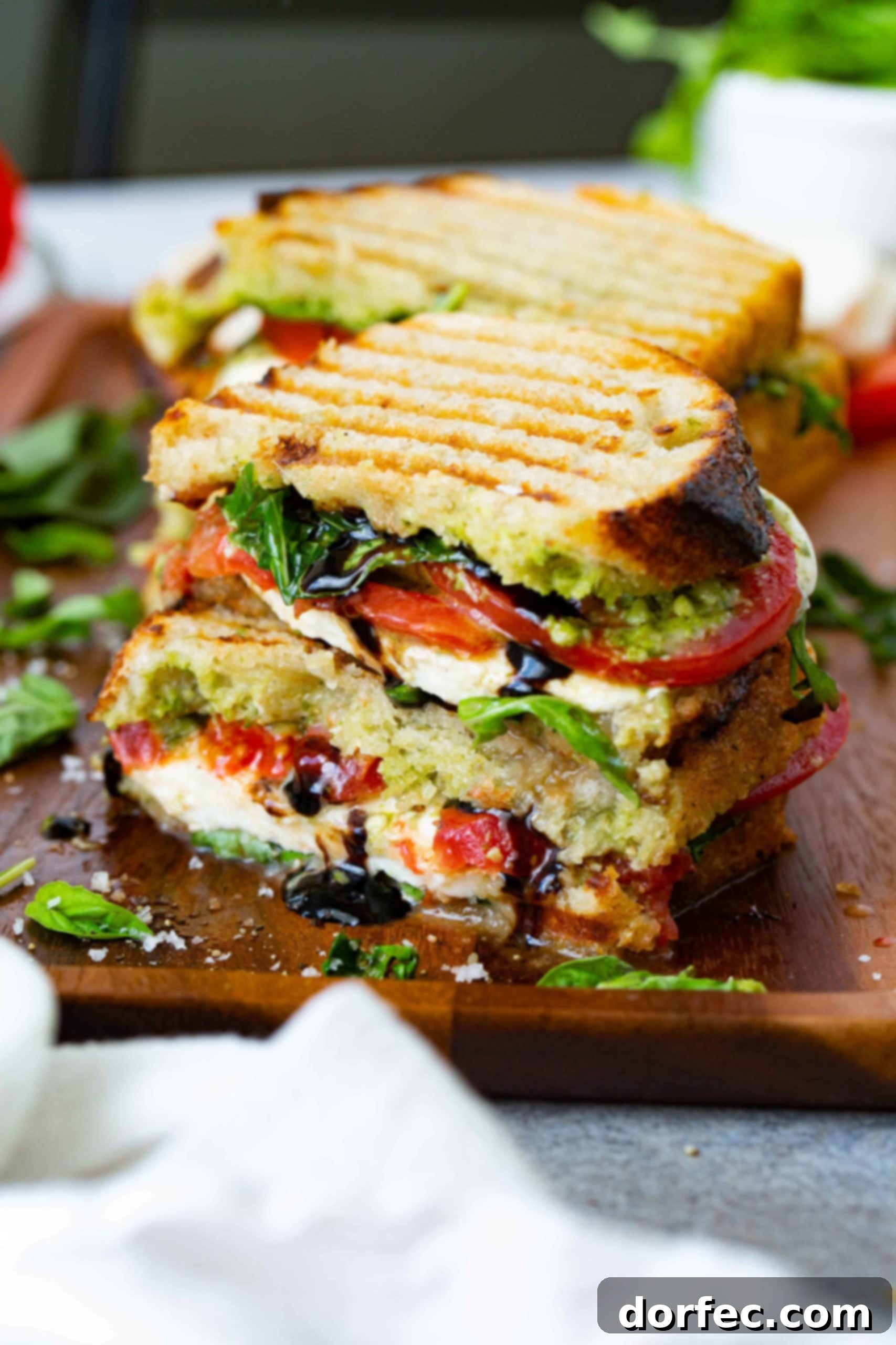 A grilled caprese panini that has been cut in half and stacked on top of each other. You can see the grill marks on the bread and the arugula, tomatoes, pesto and mozzarella cheese inside. A little balsamic glaze is oozing from the sandwich. There is another sandwich in the background, and some arugula and basil on the table.