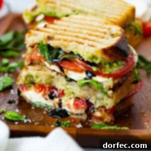 A grilled caprese panini that has been cut in half and stacked on top of each other. You can see the grill marks on the bread and the arugula, tomatoes, pesto and mozzarella cheese inside. A little balsamic glaze is oozing from the sandwich. There is another sandwich in the background, and some arugula and basil on the table.