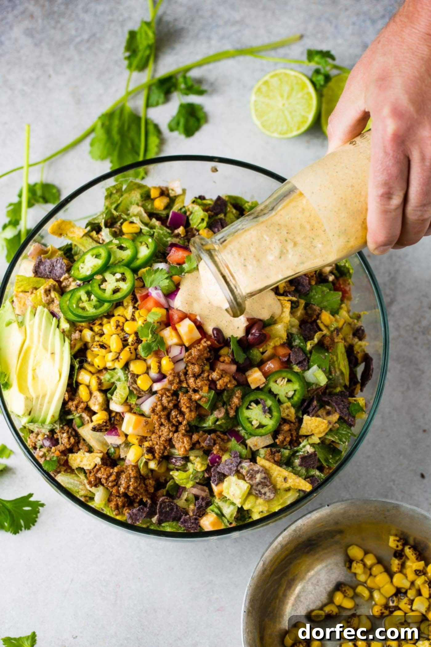 A glass bowl filled with tex-mex taco salad. The salad has avocado slices, corn, taco meat, tomatillas, tomatoes, and chips. Dressing is being poured from a glass container onto the salad.