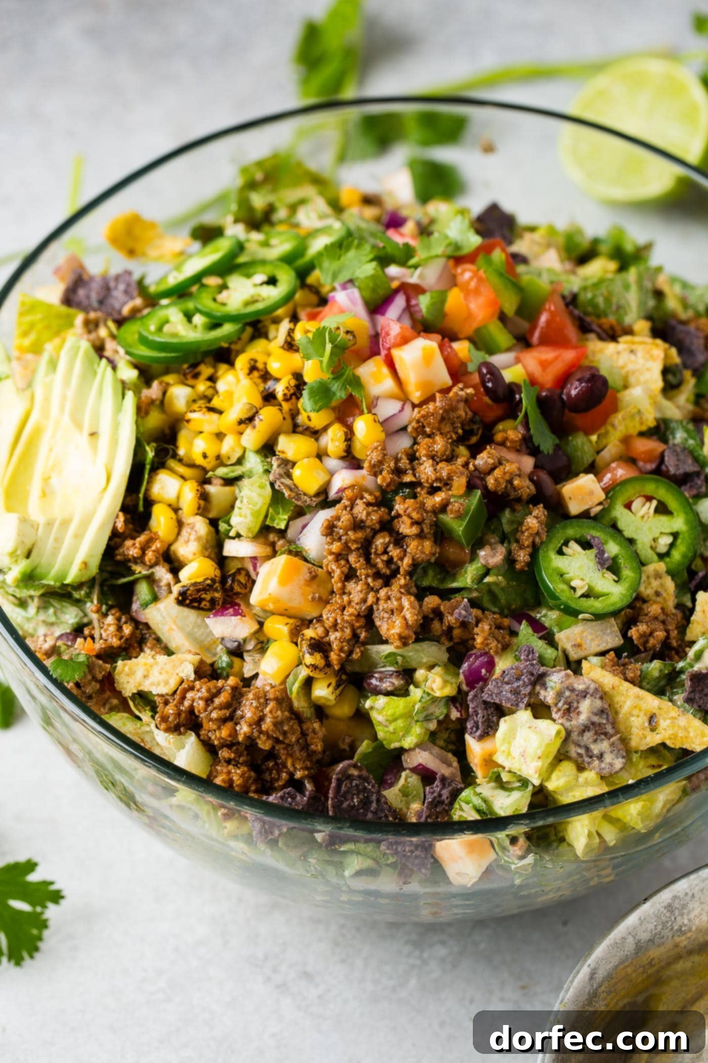 A glass bowl of tex-mex taco salad. The salad is loaded with lettuce, taco meat, corn, tomatillas, tomatoes, taco meat, and has a sliced avocado on top.