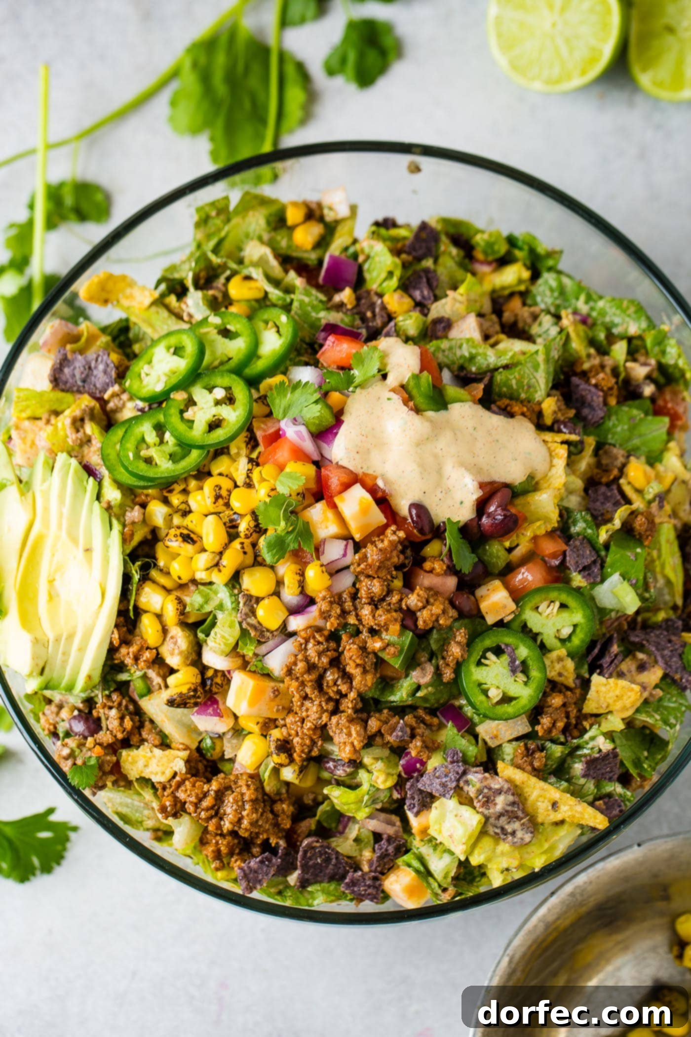 A top view of a large glass bowl of tex-mex taco salad. It is loaded with lettuce, corn, avocado, tomatillas and taco meat. There is a little dressing poured in the top part of the salad.