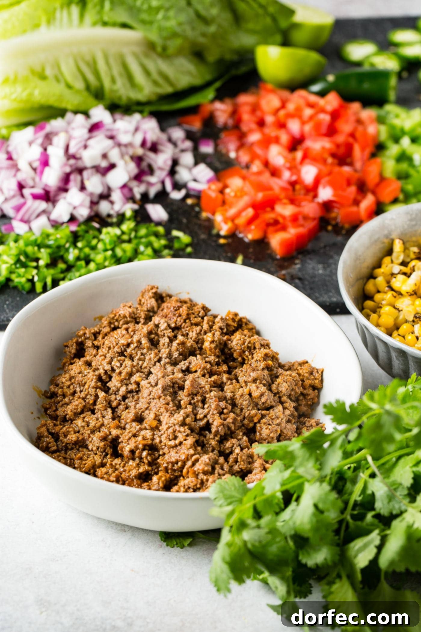 Ingredients for tex-mex taco salad. There is a bowl of taco meat, a bowl of roasted corn, cilantro, chopped green bell peppers, chopped tomatoes, and chopped red onion.