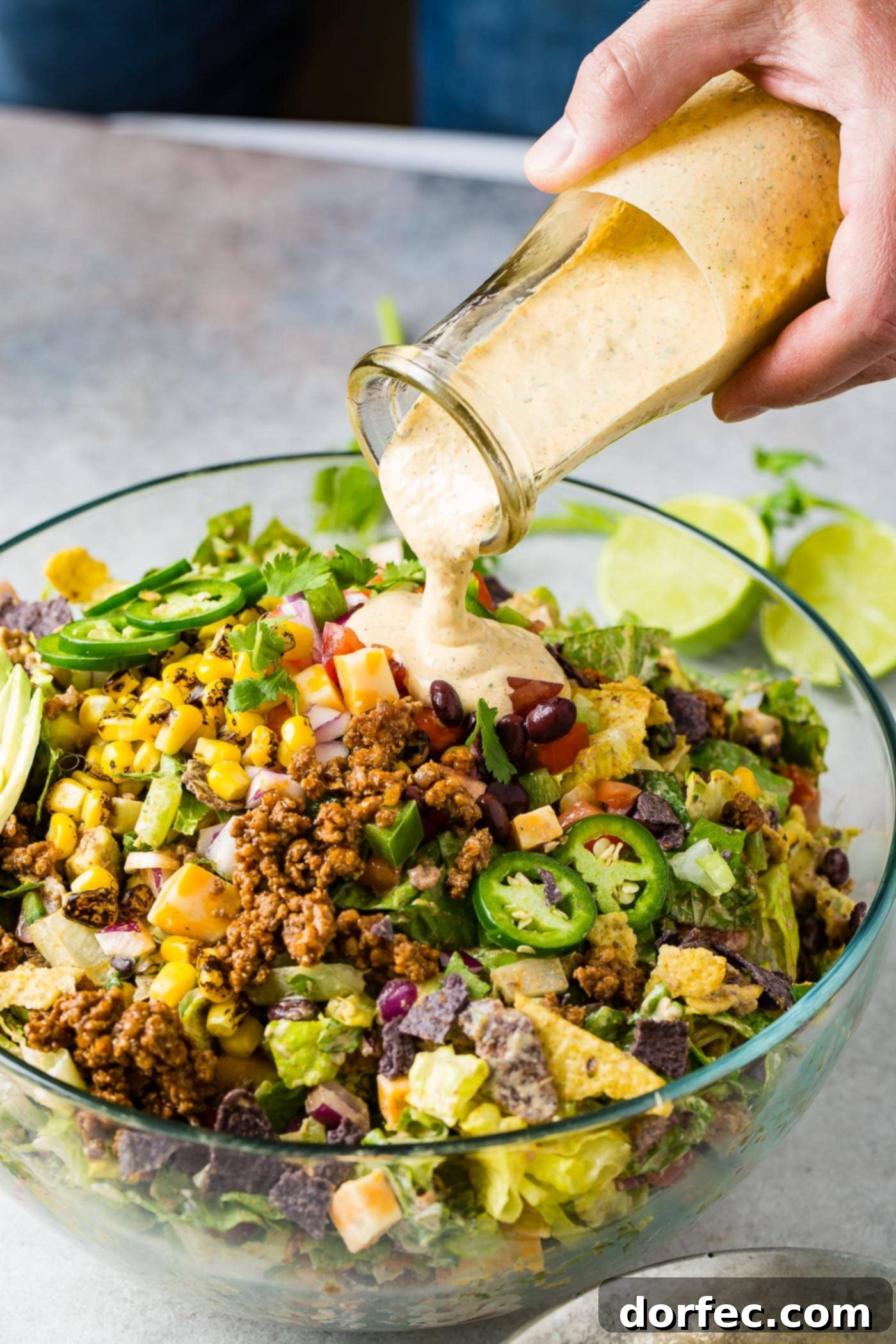 A glass bowl filled with tex-mex taco salad. A dressing is being poured over the salad. The salad is packed with corn, tomatillas, tomatoes, lettuce taco meat, and a sliced avocado is on the edge of the salad.