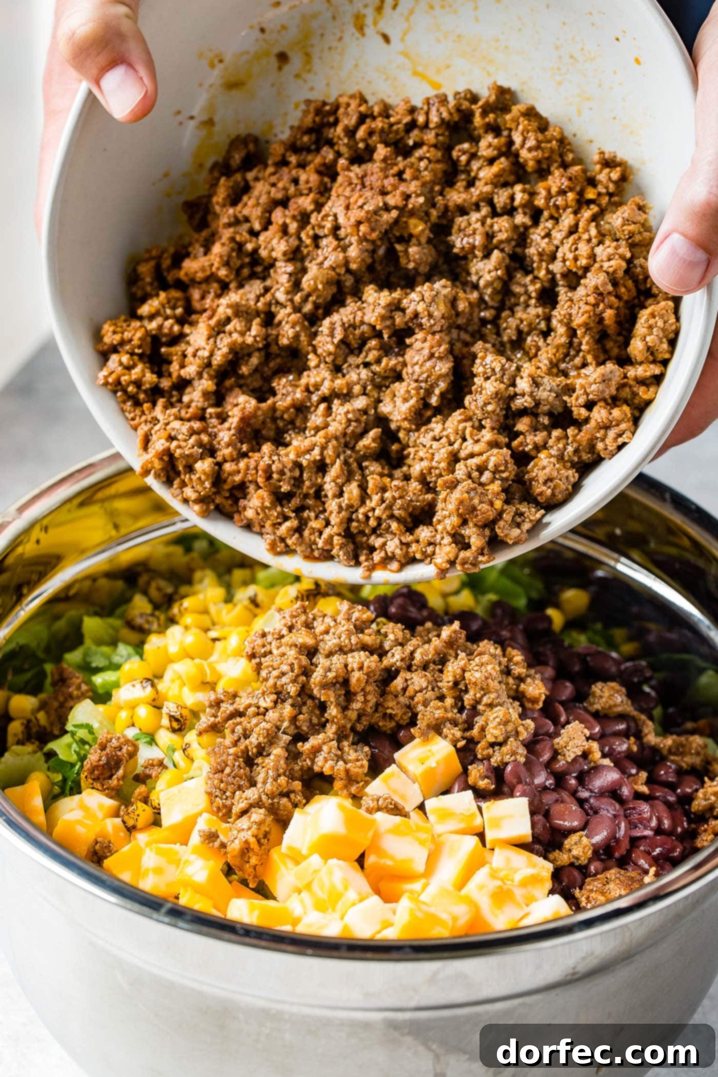 taco meat being poured into a bowl with corn, tomatillas, cheese cubes and chopped onions.