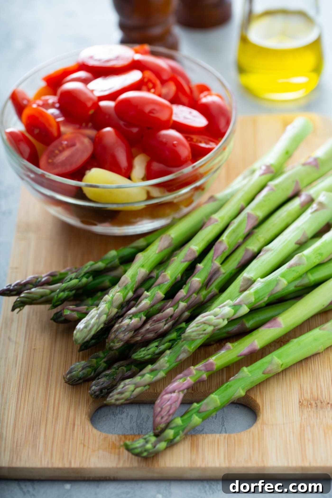 Glossy Balsamic Roasted Tomatoes and Asparagus 3 A bunch of fresh, vibrant green asparagus rests on a rustic wood cutting board. Beside it, a clear glass bowl holds an assortment of perfectly sliced red and yellow heirloom tomatoes. In the soft background, a glass container of olive oil hints at the simple, wholesome ingredients to come for this delicious roasted vegetable recipe.