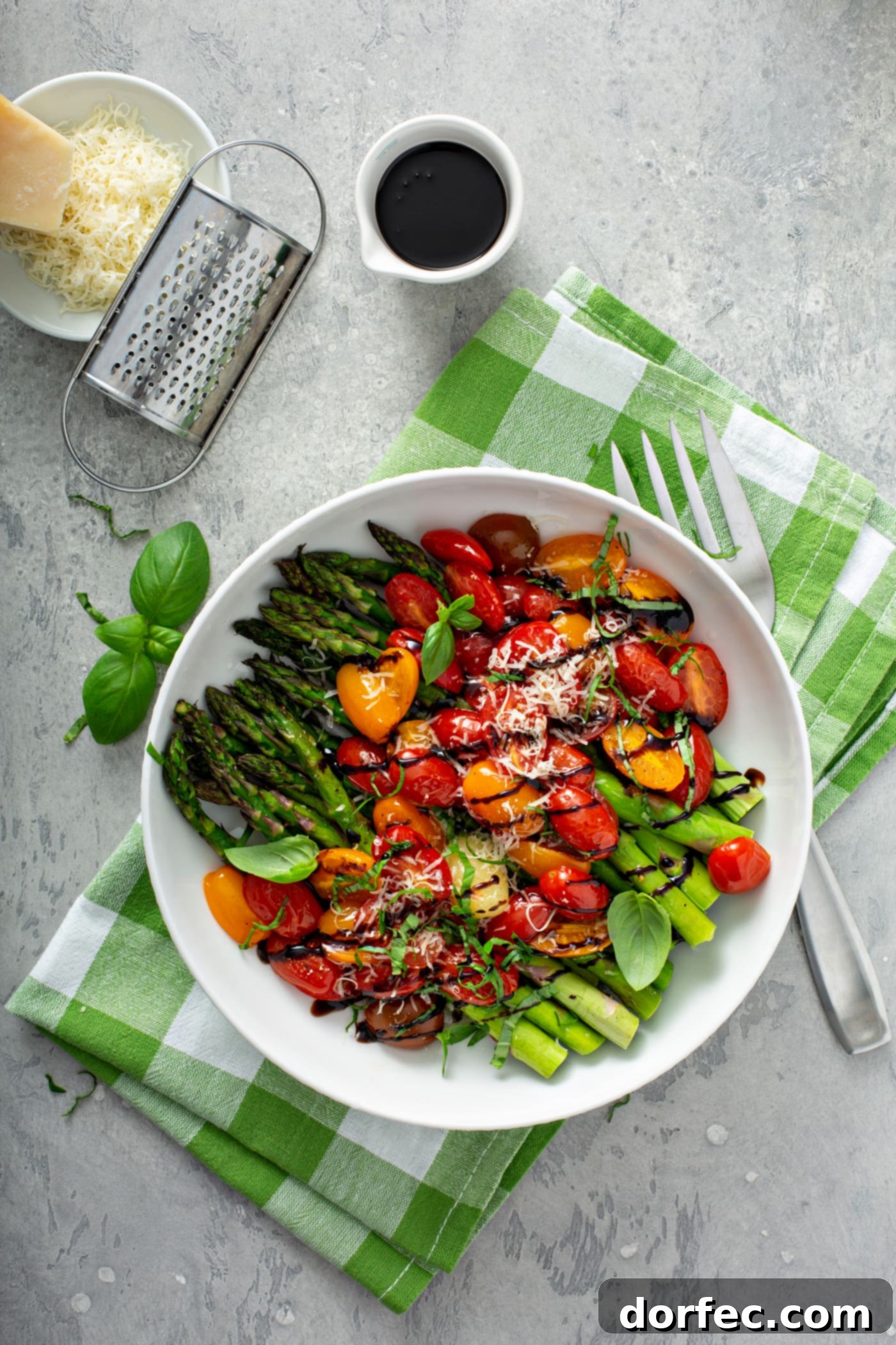 Glossy Balsamic Roasted Tomatoes and Asparagus 7 A vibrant serving dish brimming with balsamic glazed roasted tomatoes and asparagus. The bright green asparagus spears and colorful sliced red and yellow heirloom tomatoes are generously drizzled with a rich balsamic glaze and topped with a sprinkle of grated Parmesan cheese. The dish sits on a cheerful green and white checked cloth, accompanied by a fork, a cup of balsamic glaze, and a small dish of grated cheese with a grater in the background, showcasing the fresh ingredients and elegant presentation.