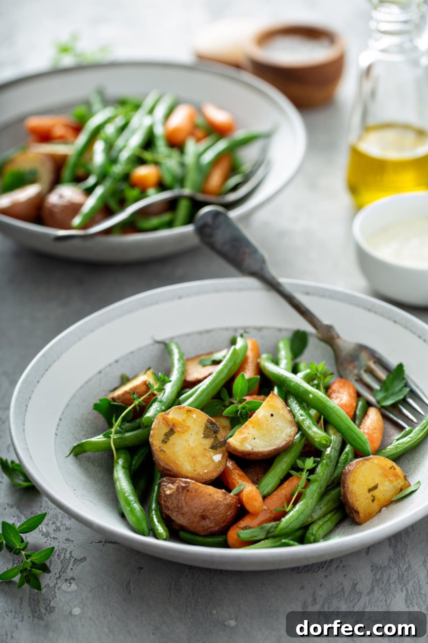 A dinner plate with vibrant garlic herb roasted vegetables, including potatoes, green beans, and carrots, with a fork resting beside them. In the background, a serving bowl filled with more roasted vegetables, a small container of salt, and a glass container of olive oil create a inviting scene.
