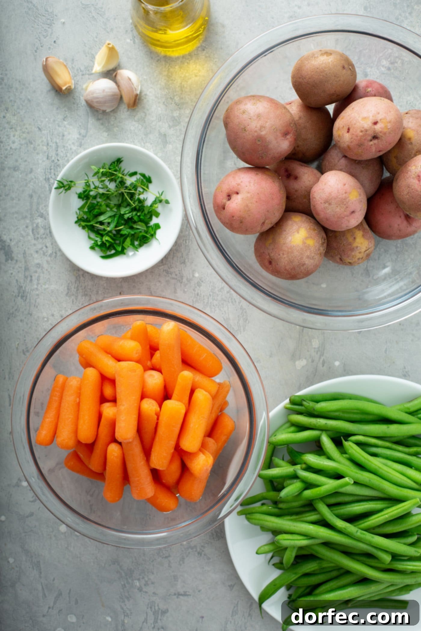 Bowls showcasing the fresh ingredients for the recipe: small red potatoes, bright orange baby carrots, crisp green beans, and a selection of vibrant green fresh herbs.