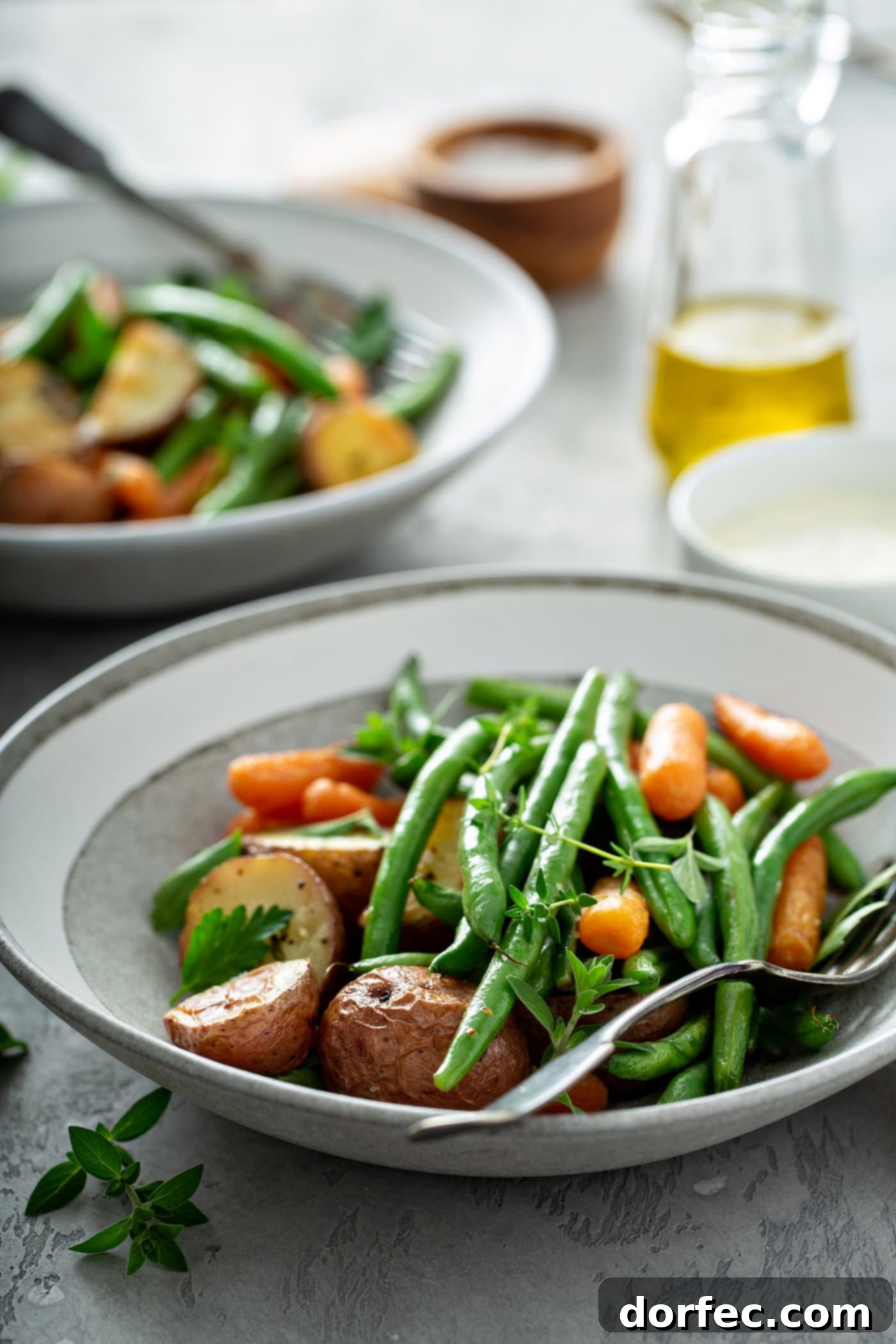 A beautifully plated dinner featuring garlic herb roasted potatoes, carrots, and green beans, with a fork resting on the plate. In the background, a serving platter brimming with more roasted vegetables, a small bowl of salt, and a glass container of olive oil complete the scene.
