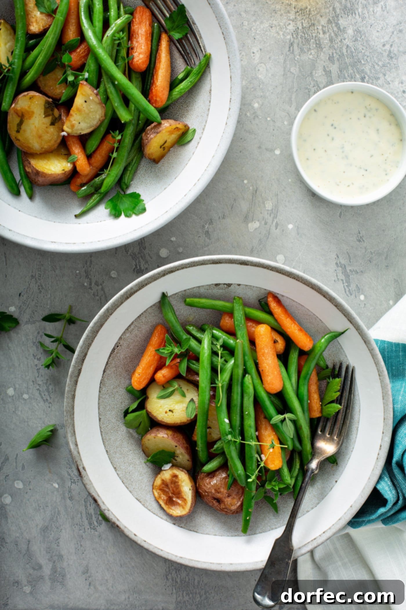Two dinner plates laden with flavorful garlic herb roasted potatoes, carrots, and green beans. Fresh parsley and thyme are artfully sprinkled over the vegetables and on the table, alongside a neatly folded napkin and a small bowl of salt in the corner.