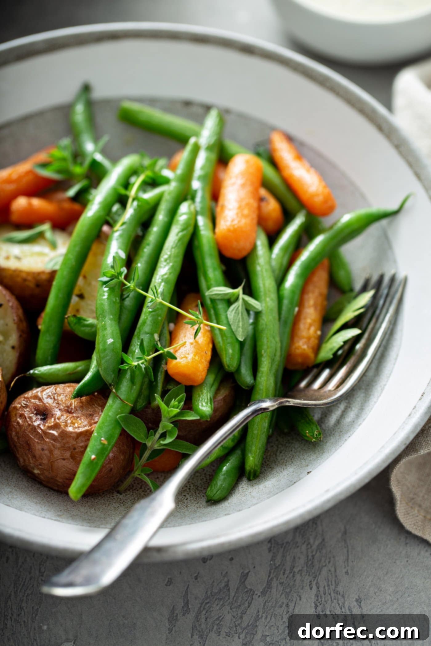 A dinner plate featuring a serving of garlic herb roasted potatoes, carrots, and green beans, garnished with fresh thyme. A fork rests neatly beside the vegetables on the plate.