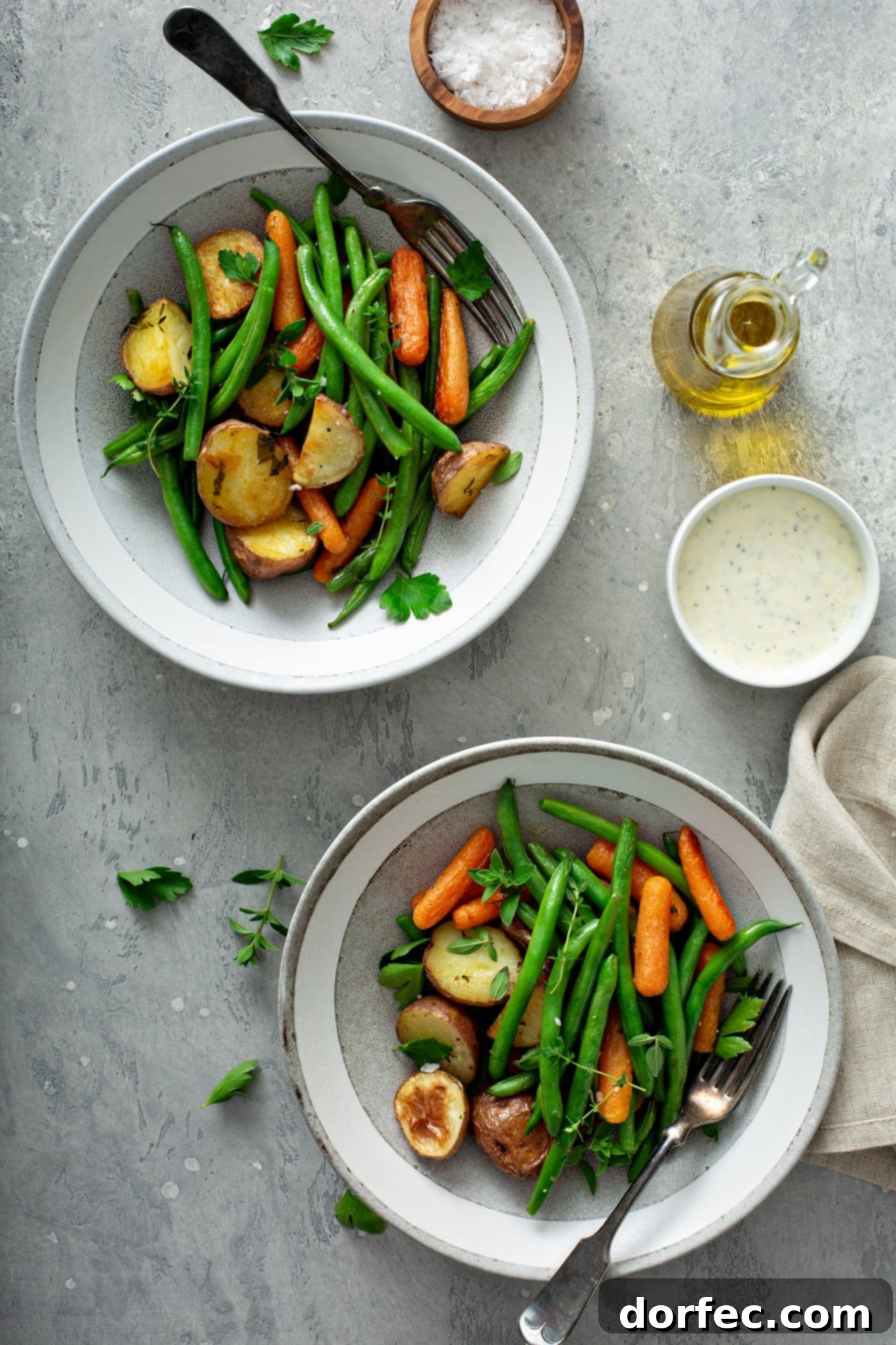 Two dinner plates, each filled with aromatic garlic herb roasted potatoes, carrots, and green beans, elegantly topped with fresh thyme and parsley. A small container of sauce, a glass container of olive oil, and a napkin are arranged on the table next to the plates.