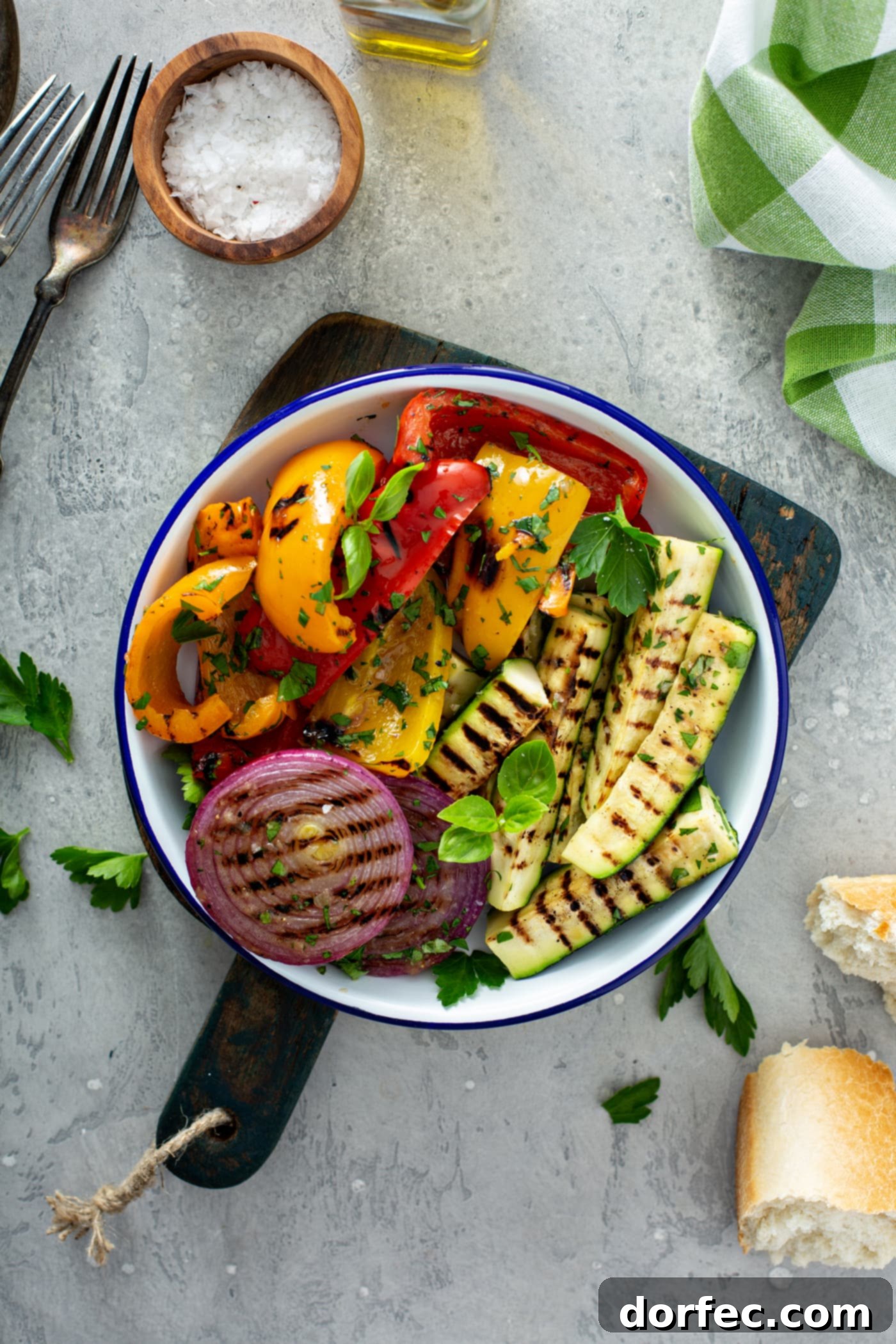 A serving plate of grilled vegetables sitting on a wooden cutting board. The grilled red onion, red and yellow bell peppers, and zucchini are topped with parsley leaves. A few parsley leaves are scattered on the table 