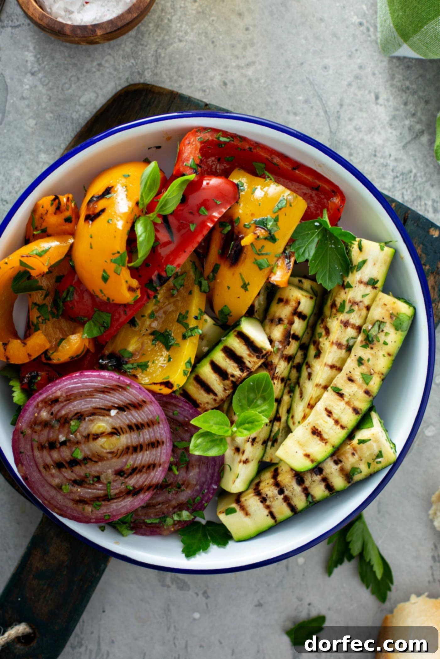 A serving dish of grilled veggies. The red onion slices, zucchini wedges and sliced red and yellow bell peppers have light brown grill marks and the veggies are sprinkled with parsley leaves.