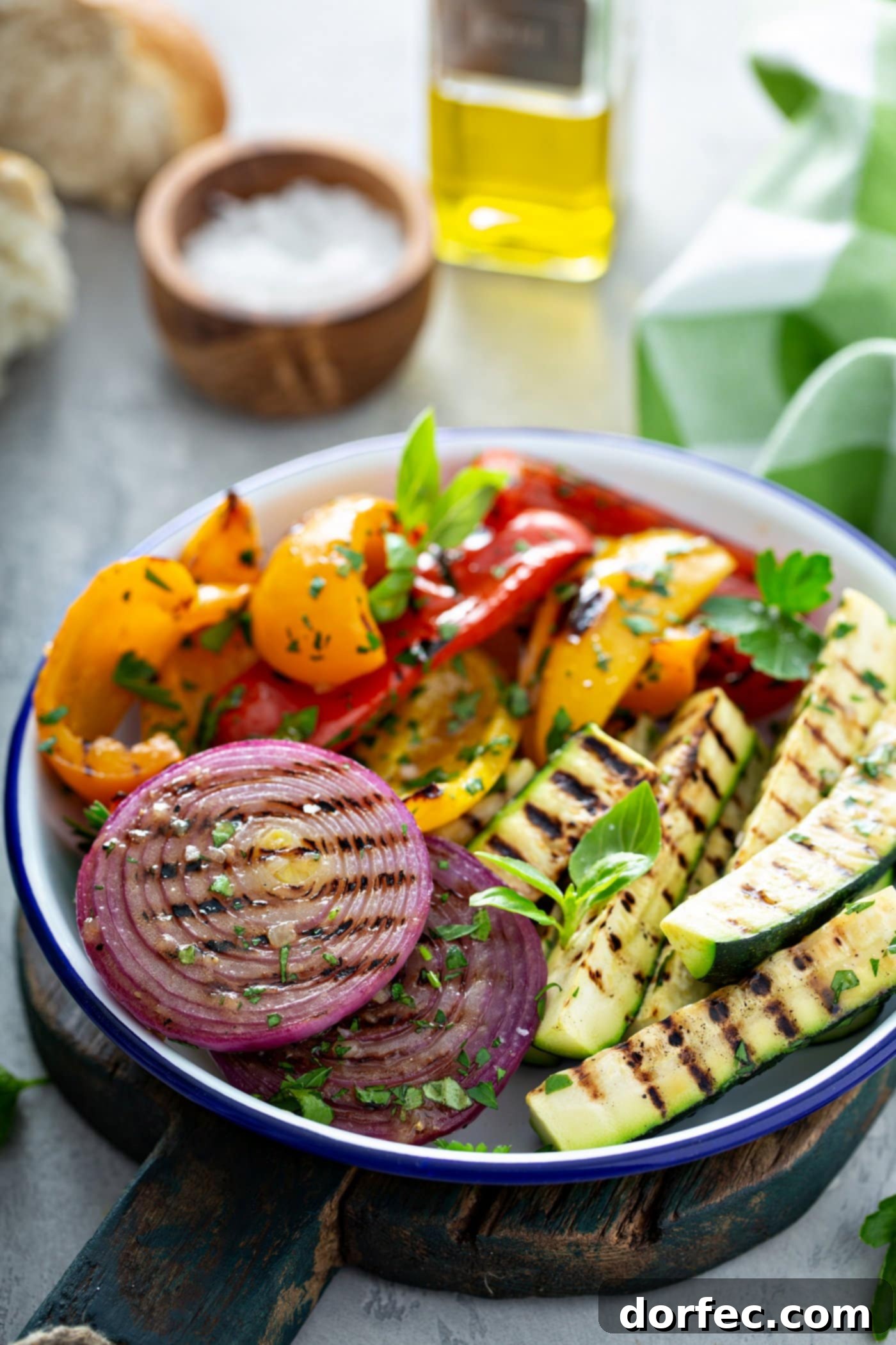 A serving dish filled with grilled veggies. There are sliced red onions, red and yellow bell peppers, and zucchini with grill marks. they are topped with a few parsley leaves. There is a wooden container of salt, a glass container of oil, and a green and white checked cloth in the background.