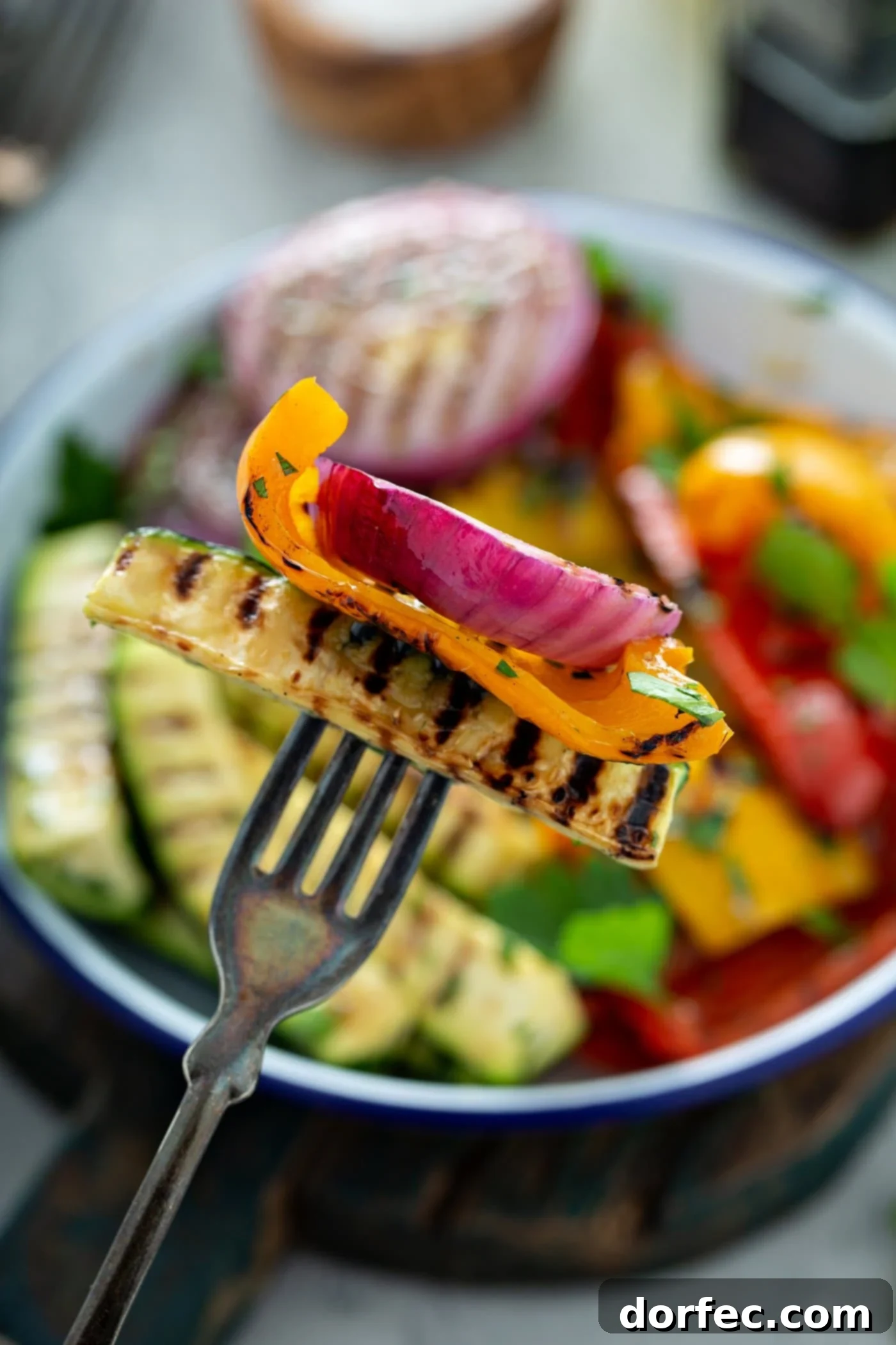 Grilled zucchini, yellow bell pepper, and red onion pierced with a fork. A serving dish of grilled veggies is in the background.