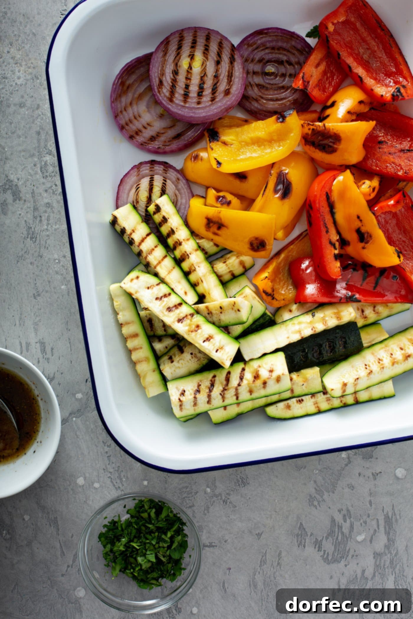 A plate of grilled veggies with a cup of balsamic glaze sitting next to the plate. The grilled zucchini wedges, slices of red onion and red and yellow bell peppers have lovely grill marks on them.