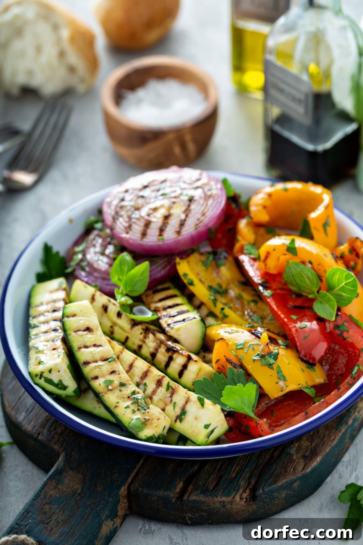 A plate of grilled vegetables. The zucchini, bell peppers, and red onions have beautiful grill marks and a few parsley leaves are scattered on top. There is a small wooden bowl of salt, a fork, and a glass cup of balsamic glaze in the background.