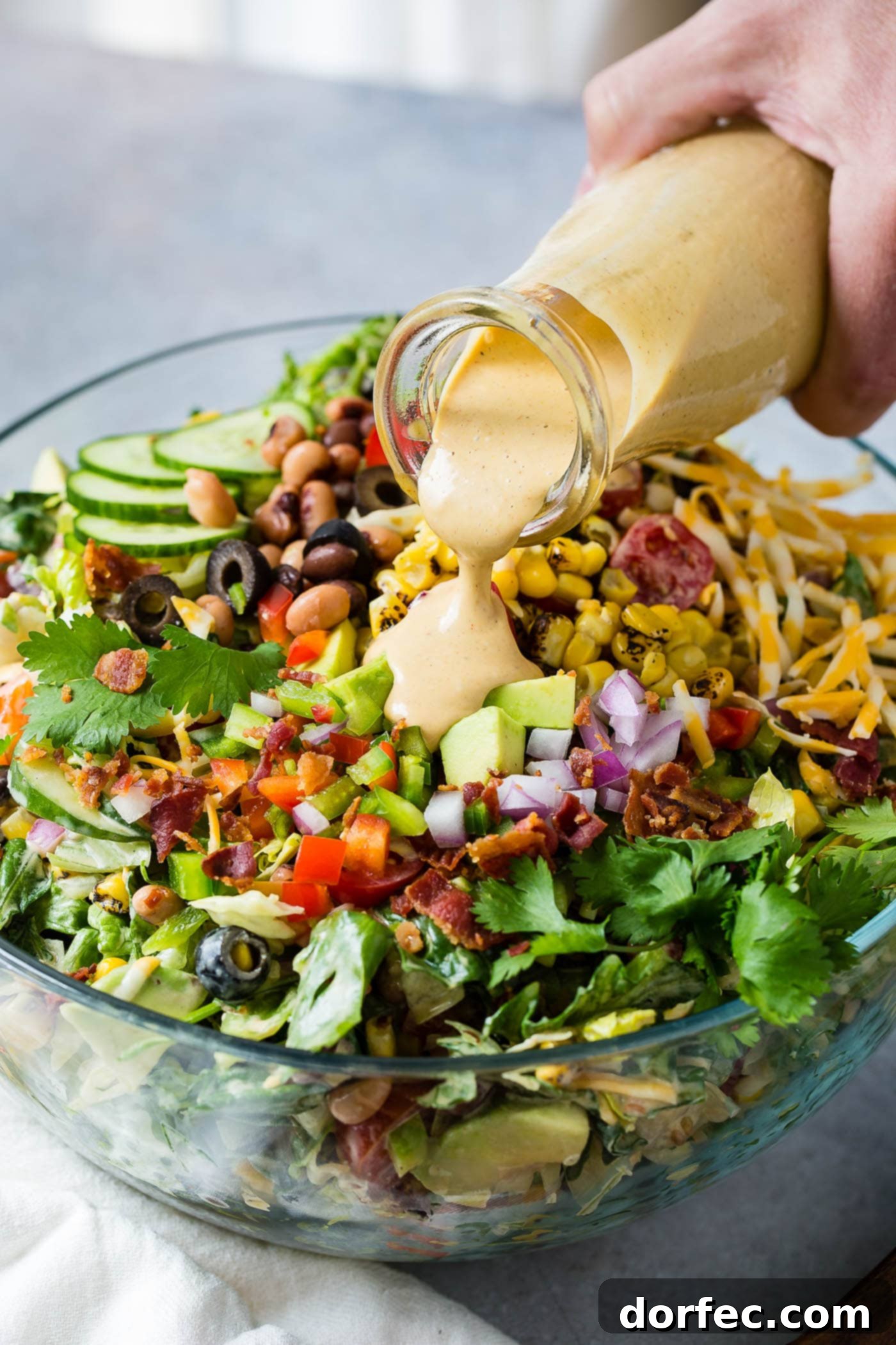 Cowboy salad in a serving bowl. A special dressing is being poured over the chopped onions, tomatoes, bacon, avocados, beans and lettuce.
