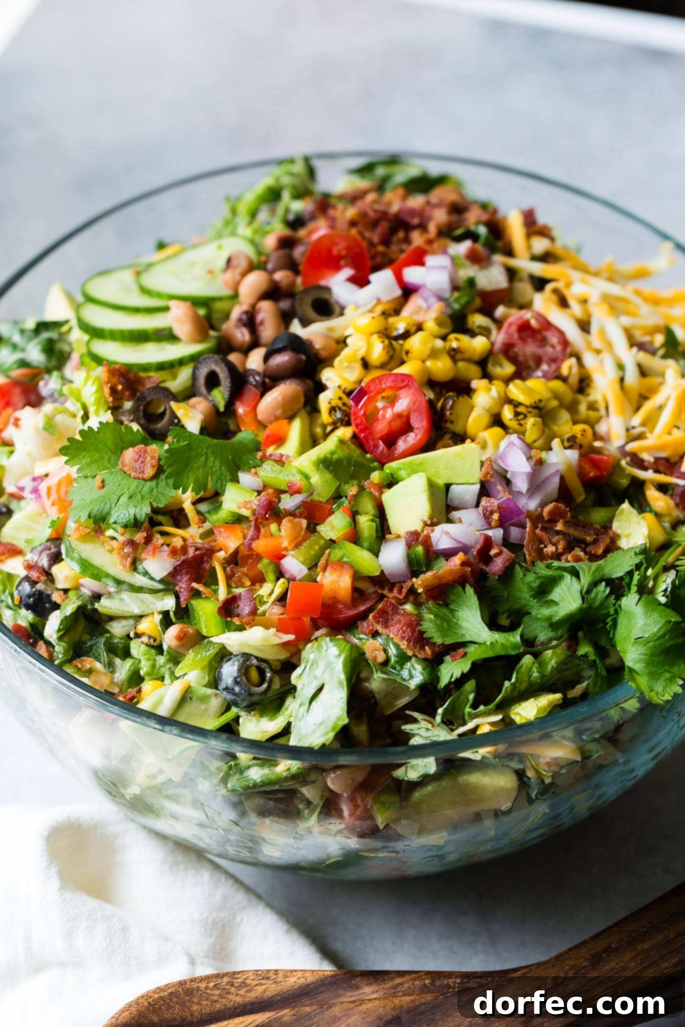 Cowboy salad that is all mixed up and ready to eat. There are sliced cucumbers, chopped tomatoes, chopped avocados, corn, beans, red onions, shredded cheese and green lettuce.