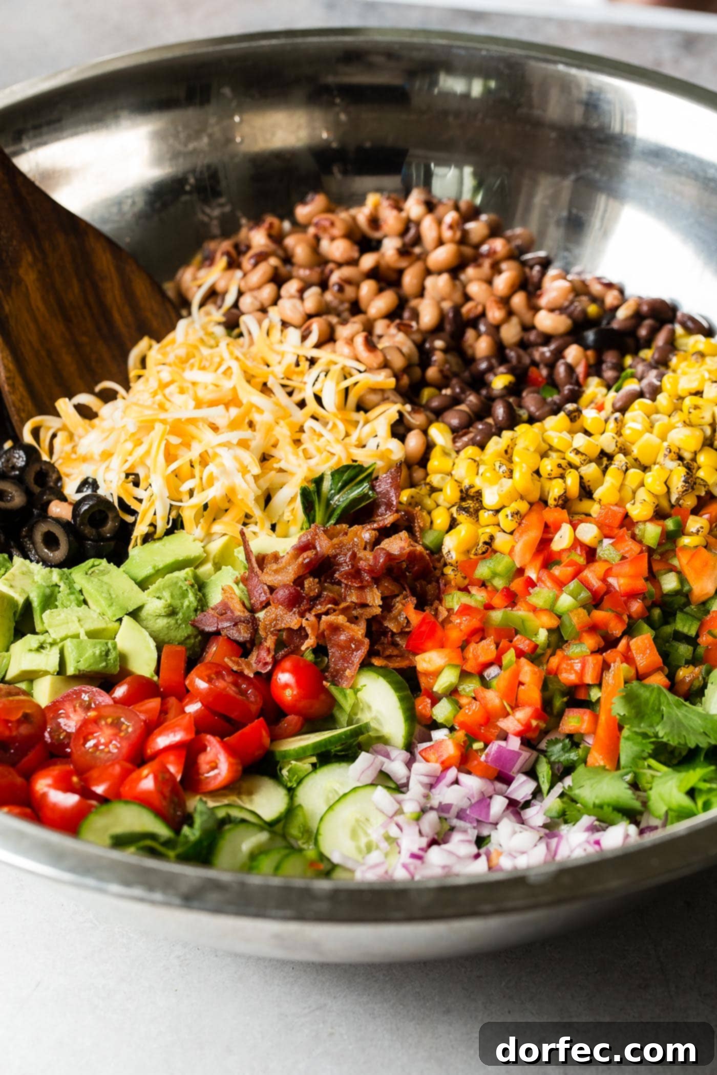 Sections of chopped tomatoes, avocados, red onion, corn, cilantro, beans, shredded cheese, olives, peppers, bacon, and peas in a glass bowl with a wooden spoon. 