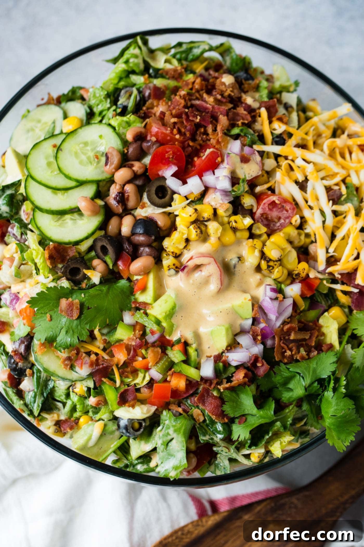 A top view of cowboy salad in a glass serving bowl. There are sliced cucumbers, sliced olives, chopped tomatoes, avocados, lettuce and green onions. It contains corn and beans too and is topped with dressing.