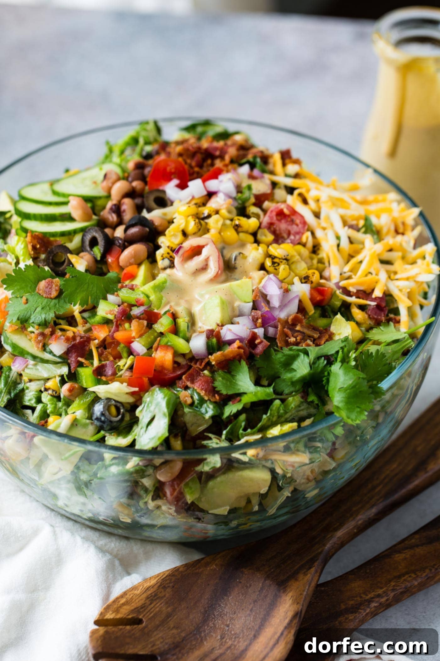 Cowboy salad in a glass serving bowl all ready to be served. It has sliced cucumbers, chopped lettuce, avocados, peppers, bacon, and olives and shredded cheese. It is topped with dressing.