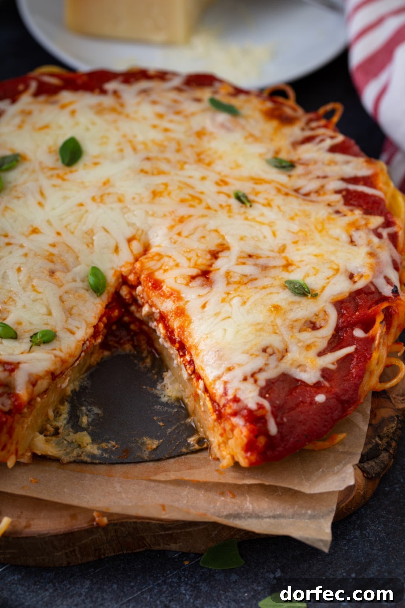 A baked spaghetti pie that has a slice removed. There are layers of spaghetti, spaghetti sauce, and melted shredded cheese. A little fresh basil is sprinkled on top.