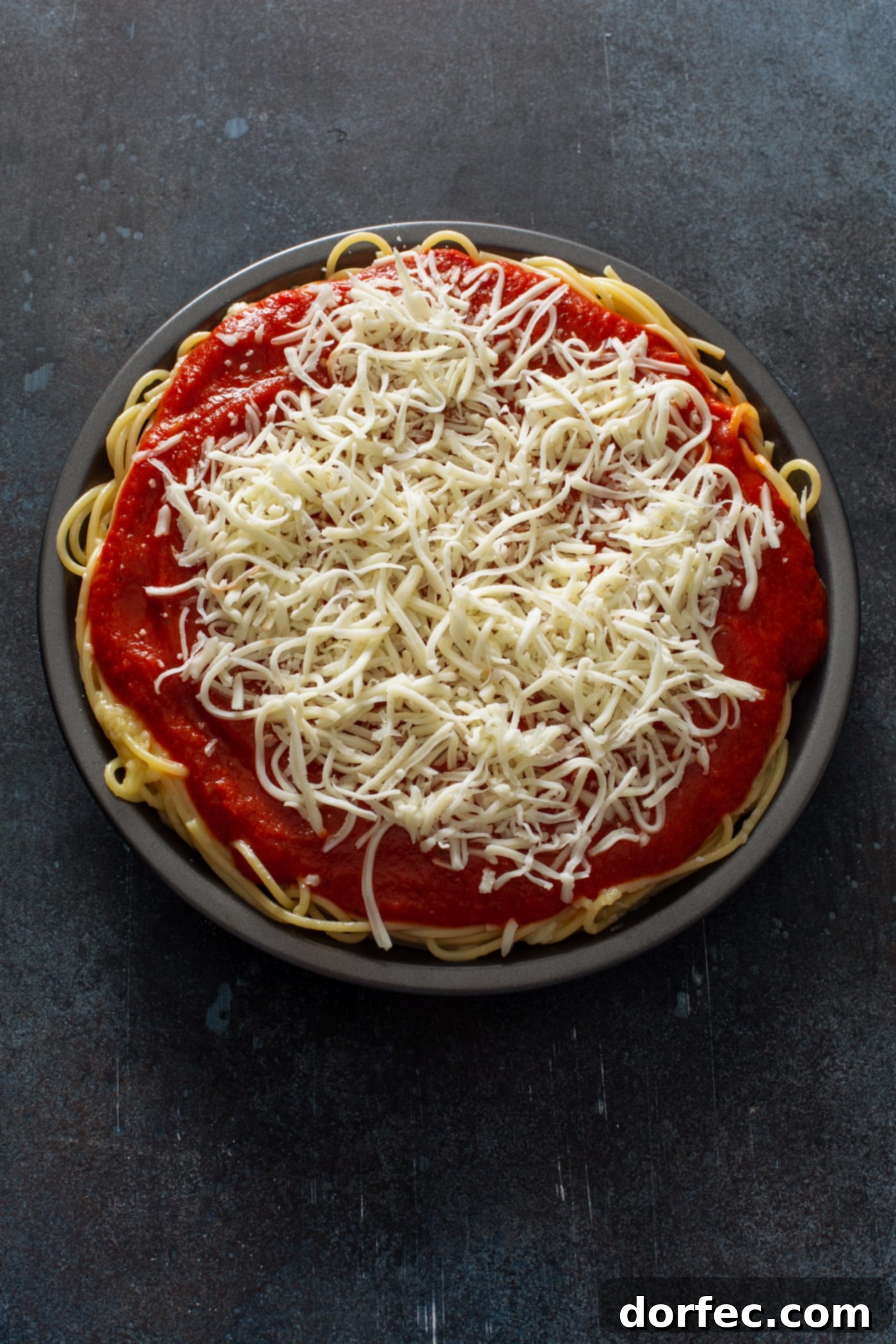 A pie dish filled with cooked spaghetti, topped with spaghetti sauce and shredded cheeses.