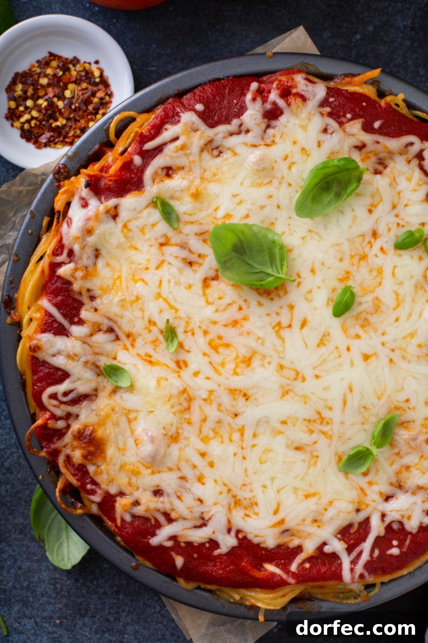 A top view of baked spaghetti pie. There is spaghetti pasta, spaghetti sauce and melted cheeses with a few basil leaves on top. A small bowl of dried pepper flakes is behind the pie.