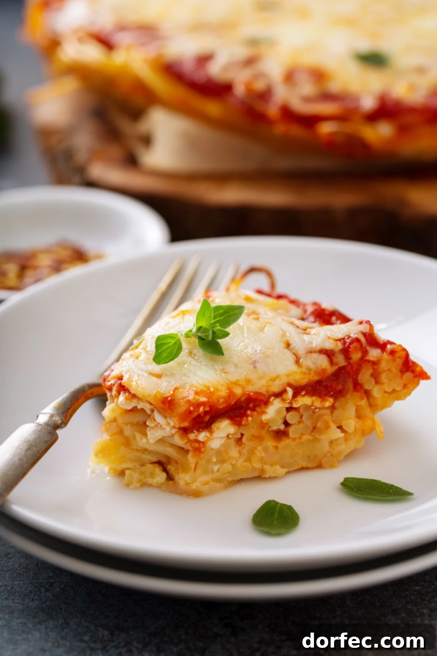 A slice of baked spaghetti pie on a white dinner plate. A fork is next to the spaghetti pie and a few basil leaves are scattered on the pie and the plate. A small container of red pepper flakes and the rest of the spaghetti pie are in the background.