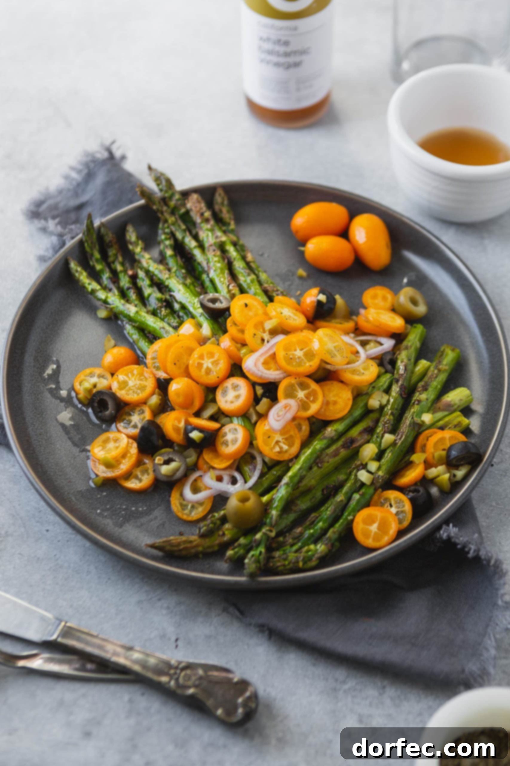 A colorful arrangement of fresh ingredients for the roasted asparagus and kumquats recipe, including bright green asparagus spears, whole orange kumquats, a bottle of olive oil, and a small bowl of olives, ready for preparation.