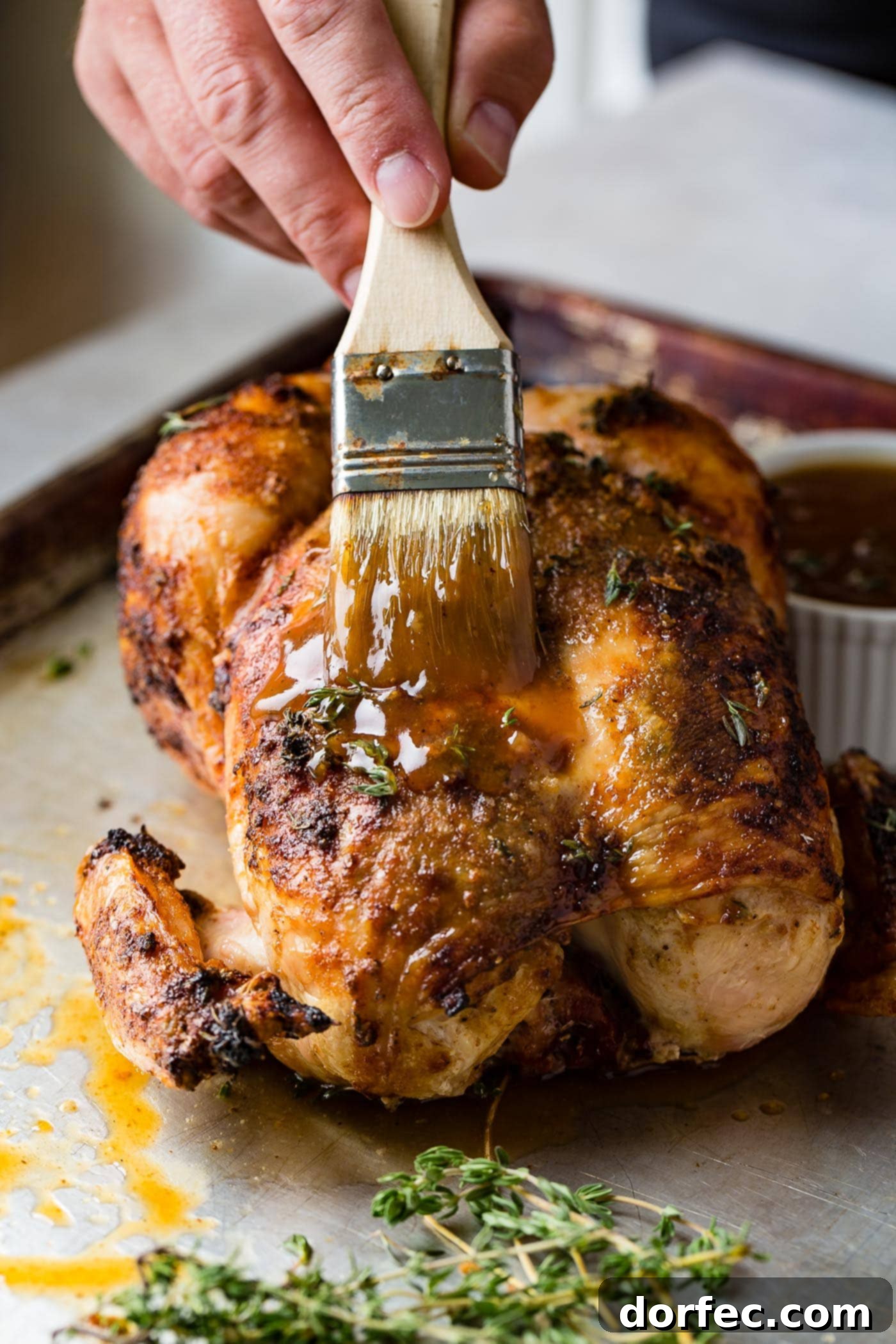 A succulent smoked whole chicken is being brushed with a savory glaze, enhancing its golden-brown crust. A small bowl of gravy and a dinner plate are visible in the background, completing the inviting scene.