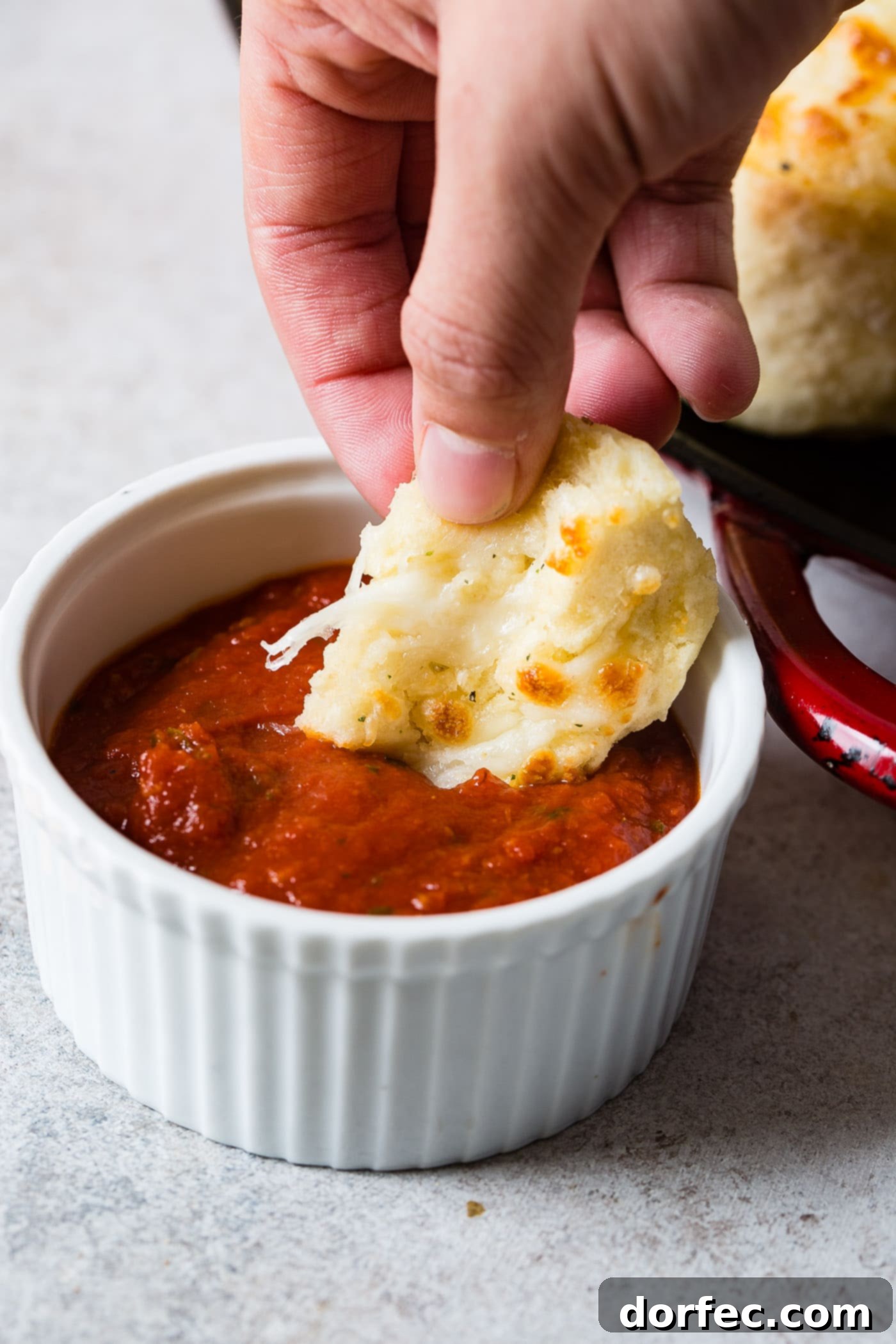 A piece of cheese doodle cheesy bread being dipped in a a small white bowl of marinara sauce. A tray of cheese doodles sits next to the sauce.