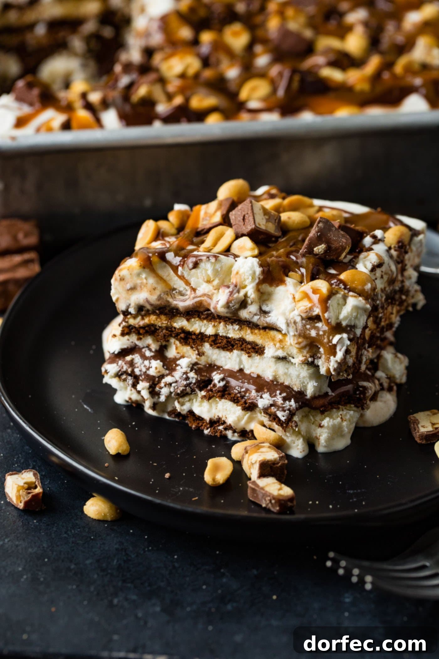 A beautifully layered slice of Snickers Ice Cream Cake on a brown dessert plate, showcasing distinct strata of chocolate cookie, vanilla ice cream, rich hot fudge, golden caramel, whipped cream, and chopped Snickers bars, all generously sprinkled with salted peanuts. In the blurred background, a full pan of the irresistible cake awaits.