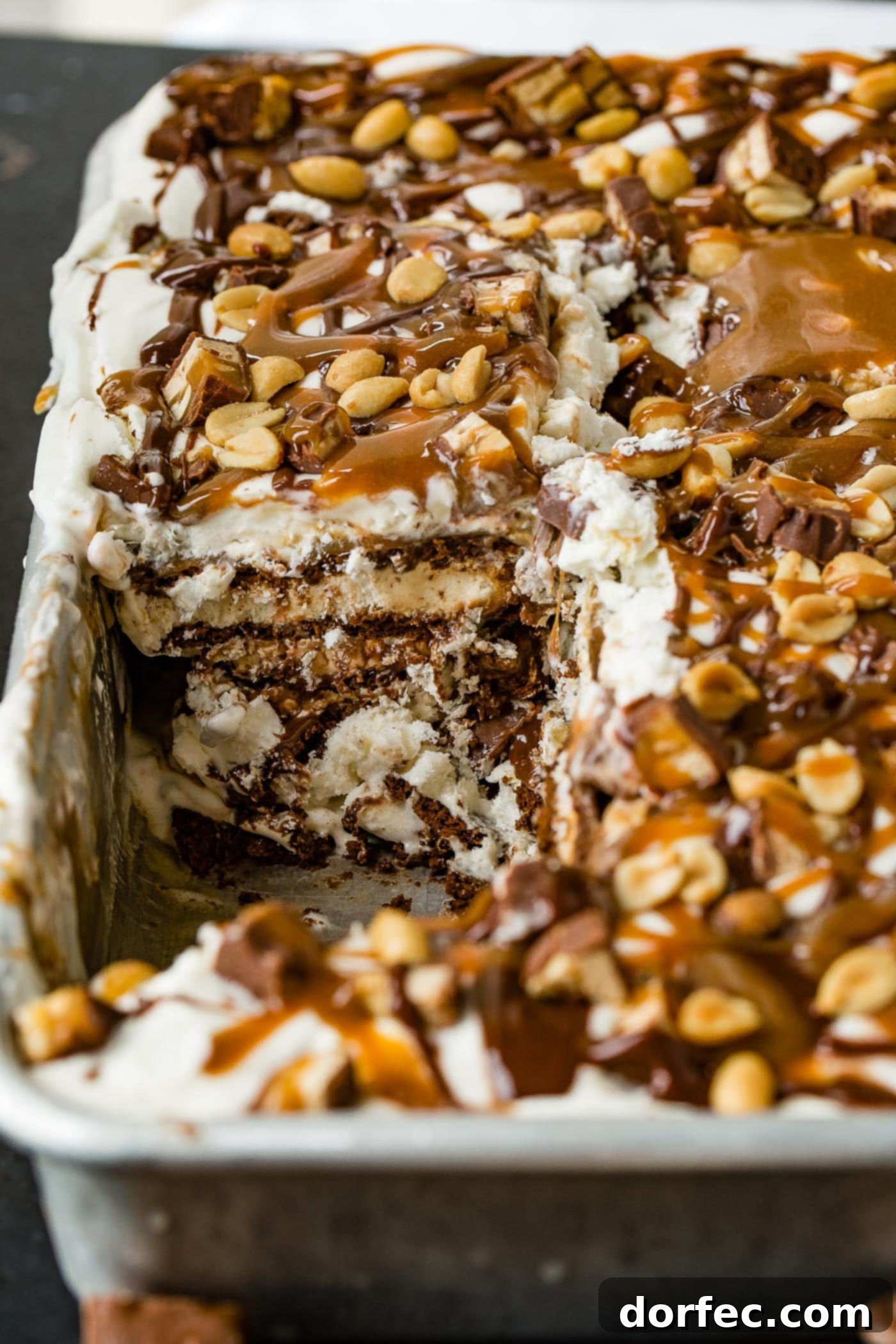 A delectable Snickers Ice Cream Cake in a 9x13 inch pan, with a piece already removed, revealing the exquisite layers. You can clearly see the chocolate cookie of the ice cream sandwiches, creamy ice cream, thick hot fudge, gooey caramel sauce, light whipped cream, and scattered chunks of Snickers candy bars. The top is artfully drizzled with more hot fudge and caramel, and generously topped with salted peanuts.