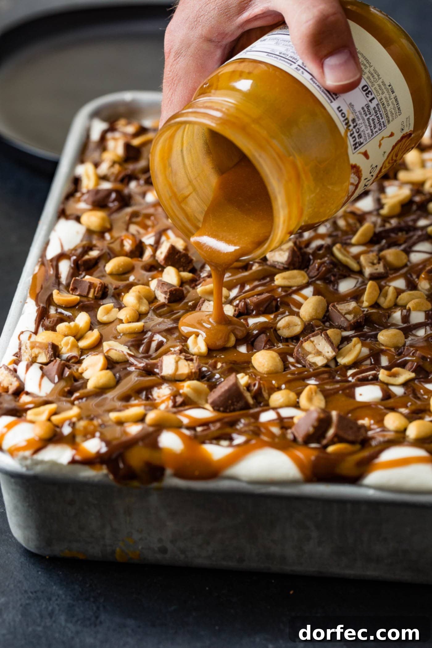 A close-up shot of rich, melted caramel topping being poured from a glass jar, elegantly drizzling over the pristine white whipped cream surface of a Snickers Ice Cream Cake. The top of the cake is already adorned with more hot fudge drizzles and scattered salted peanuts, creating a tempting visual of sweet and salty indulgence.