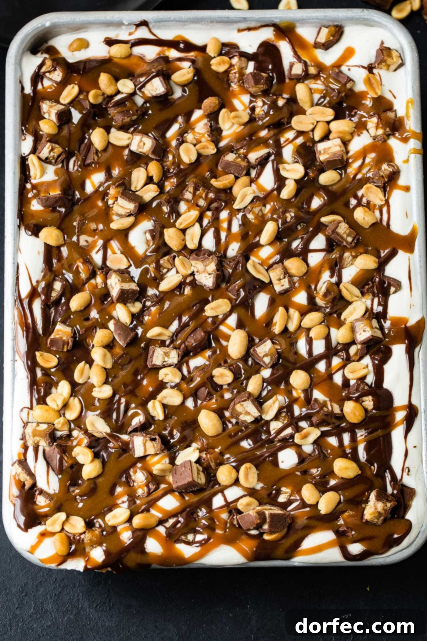 A stunning top-down view of a completed Snickers Ice Cream Cake in its pan, ready for serving. The surface is generously blanketed with fluffy white whipped cream, crisscrossed with decadent drizzles of hot fudge and golden caramel sauce, and beautifully scattered with chopped Snickers bars and crunchy salted peanuts. A truly enticing no-bake dessert.