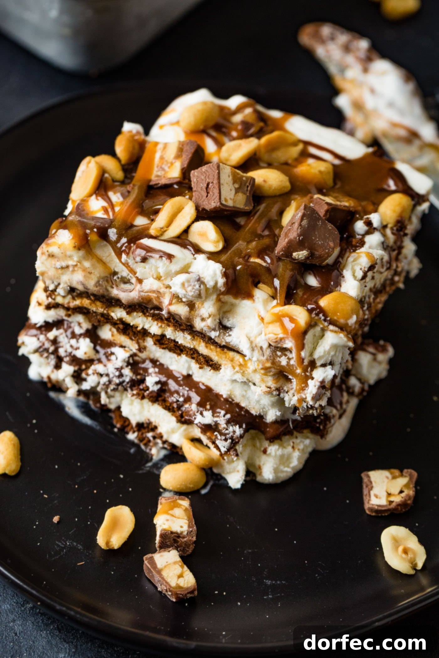 A perfect slice of Snickers Ice Cream Cake presented on a white dessert plate, adorned with a delicate chocolate swirl. The cross-section reveals beautifully defined layers of dark chocolate cookie, creamy ice cream, rich hot fudge, gooey caramel sauce, and fluffy whipped cream. The top is generously garnished with hot fudge, caramel drizzles, and a sprinkle of salted peanuts, with extra Snickers chunks and peanuts artfully scattered around the plate.