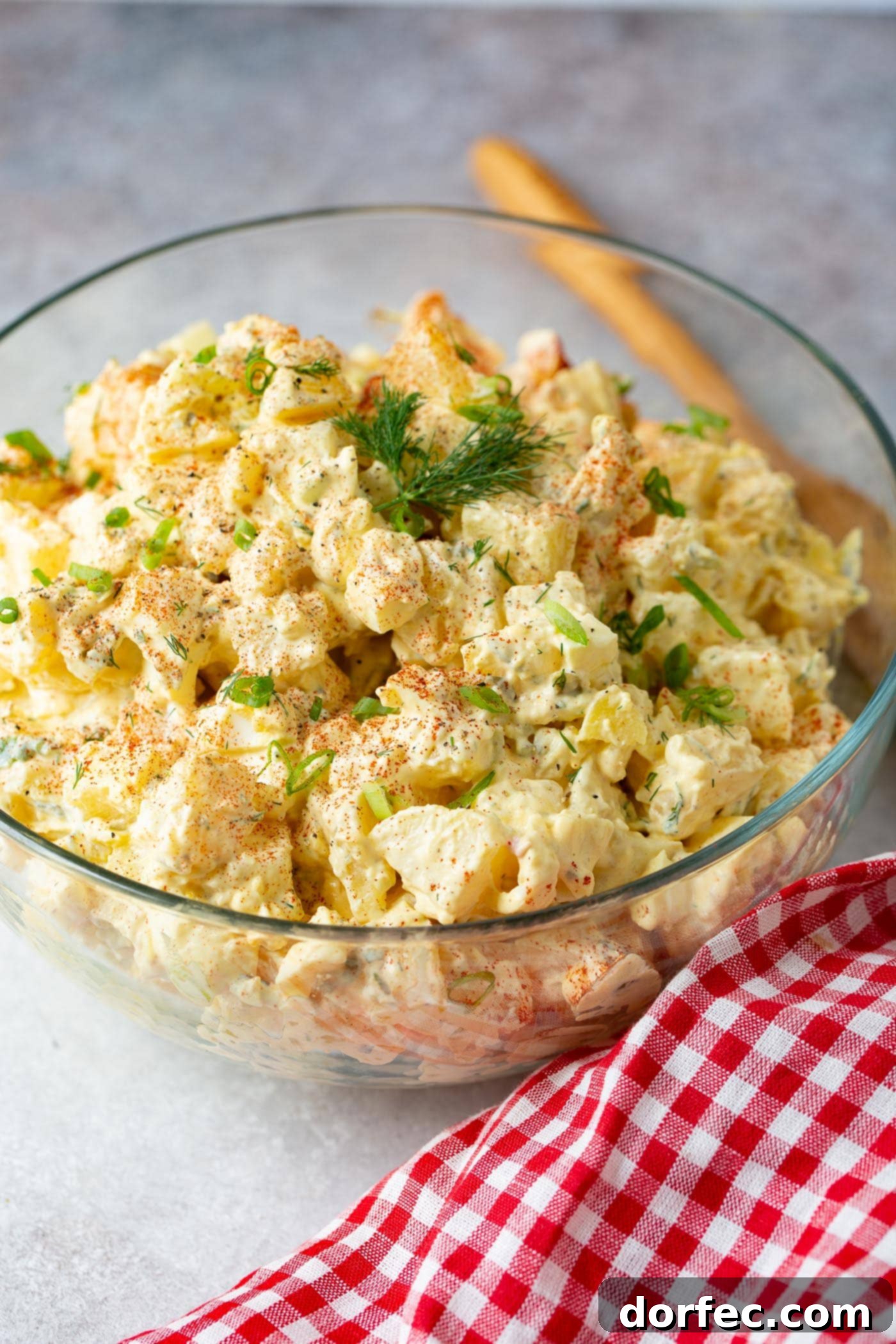 The Only Potato Salad Recipe You'll Ever Need 2 A glass serving bowl filled with a BBQ side! There are chunks of chopped eggs, fresh dill, and it is sprinkled with paprika and fresh parsley leaves. The bowl is sitting on a table next to a red checked napkin. A wooden spoon is also laying on the table.