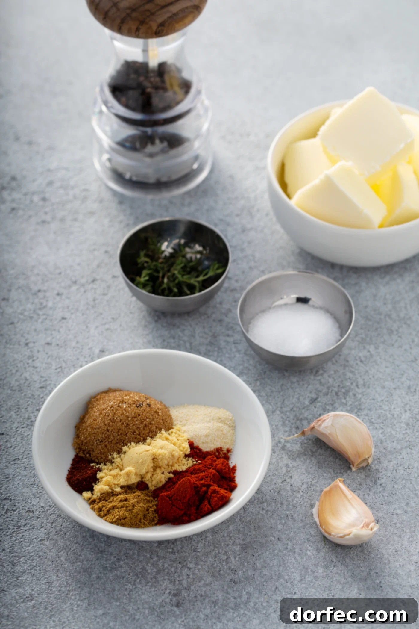 Small containers holding cubed butter, salt, and pepper are arranged next to a bowl containing individual mounds of brown sugar, paprika, onion powder, cayenne pepper, and ground mustard, along with two cloves of garlic.