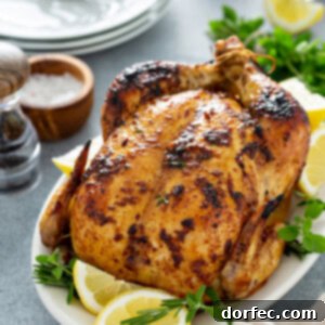 A whole roasted chicken on a white platter with lemon wedges and fresh thyme. On the table next to the chicken is a pepper shaker, salt in a wooden bowl, a stack of 3 dinner plates, and fresh thyme.