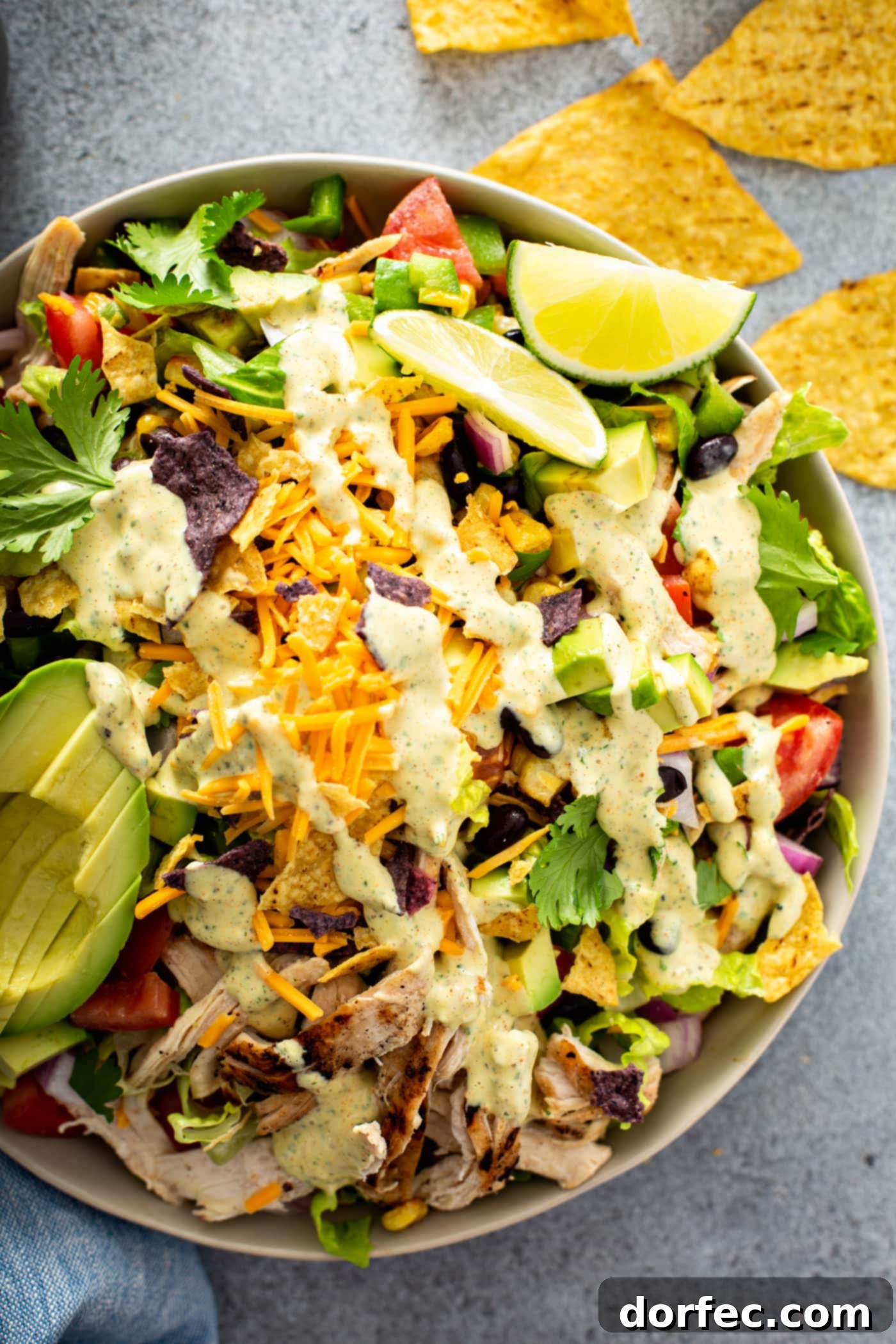 Simple Chicken Taco Salad A large bowl of chicken taco salad. There is shredded chicken, cheese, cilantro, beans, corn, tomatoes, red onions, peppers and sliced avocados. There are 2 lime wedges next to the salad and chips and dressing in the background.